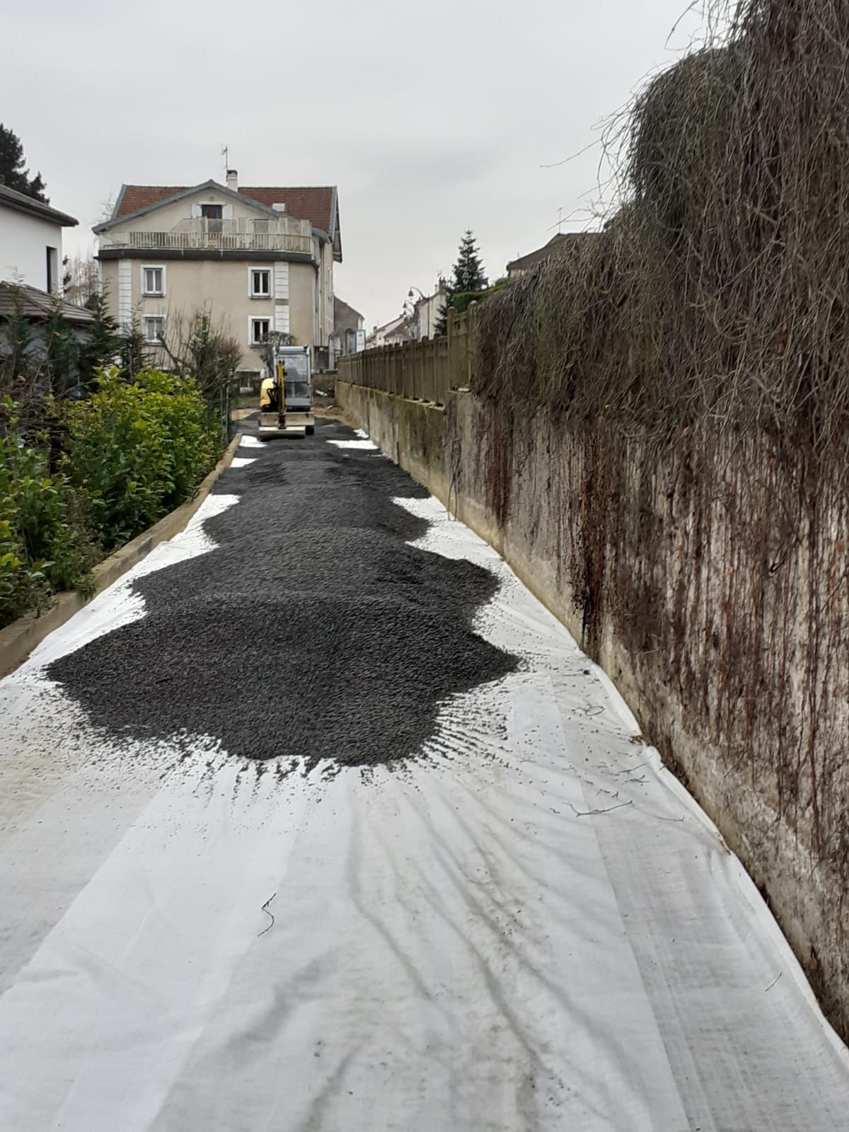 Chantier : gravier noir sur toile blanche ; excavatrice au loin, à côté d'un mur recouvert de vigne et de bâtiments.