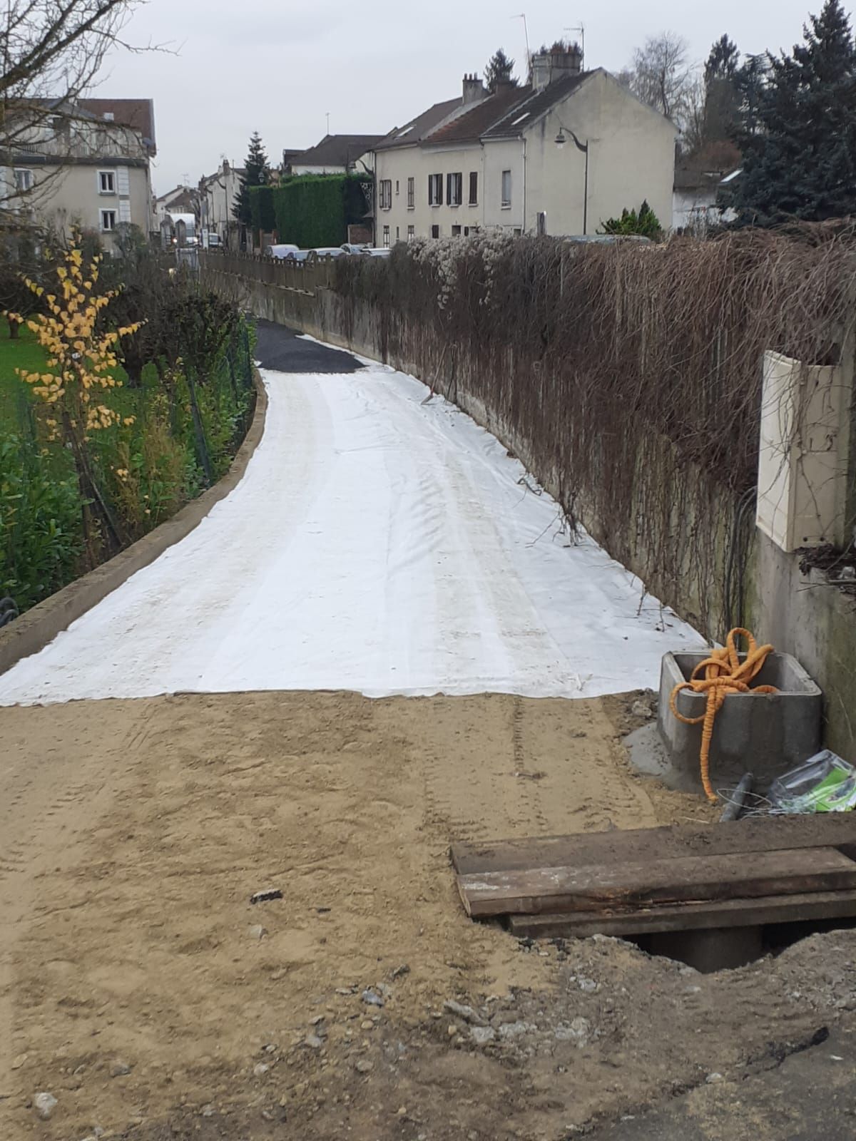Une bâche blanche recouvre un chemin longeant une haie, menant à des maisons dans une ville.