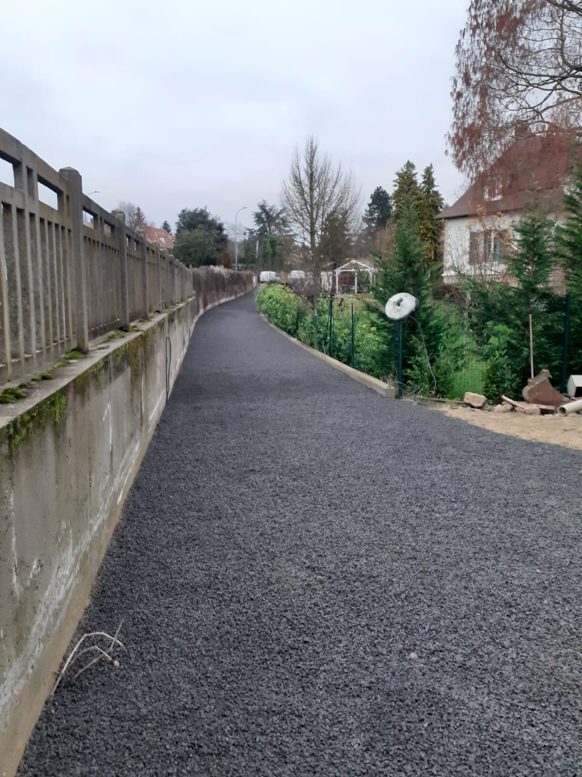 Allée pavée entre un mur de béton et de la verdure, menant vers des maisons sous un ciel couvert.
