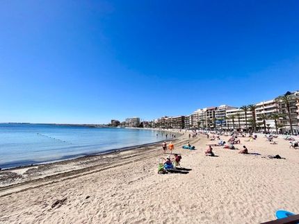Una playa llena de gente en un día soleado con edificios al fondo.