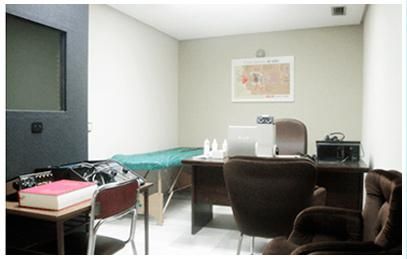 Pequeña sala de examen con escritorio, sillas, mesa de exploración y mapa mural.