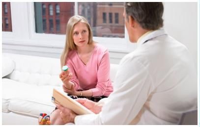 Mujer con camisa rosa hablando con un médico en un consultorio luminoso, sosteniendo un frasco de medicina.