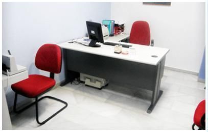 Pequeña oficina con un escritorio blanco, un monitor de ordenador y dos sillas rojas sobre un suelo claro.