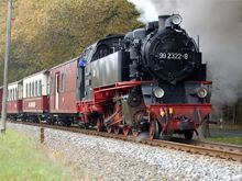 Eine schwarze Dampflokomotive zieht rote Personenwagen auf Gleisen, Rauch steigt in einer ländlichen Umgebung auf.