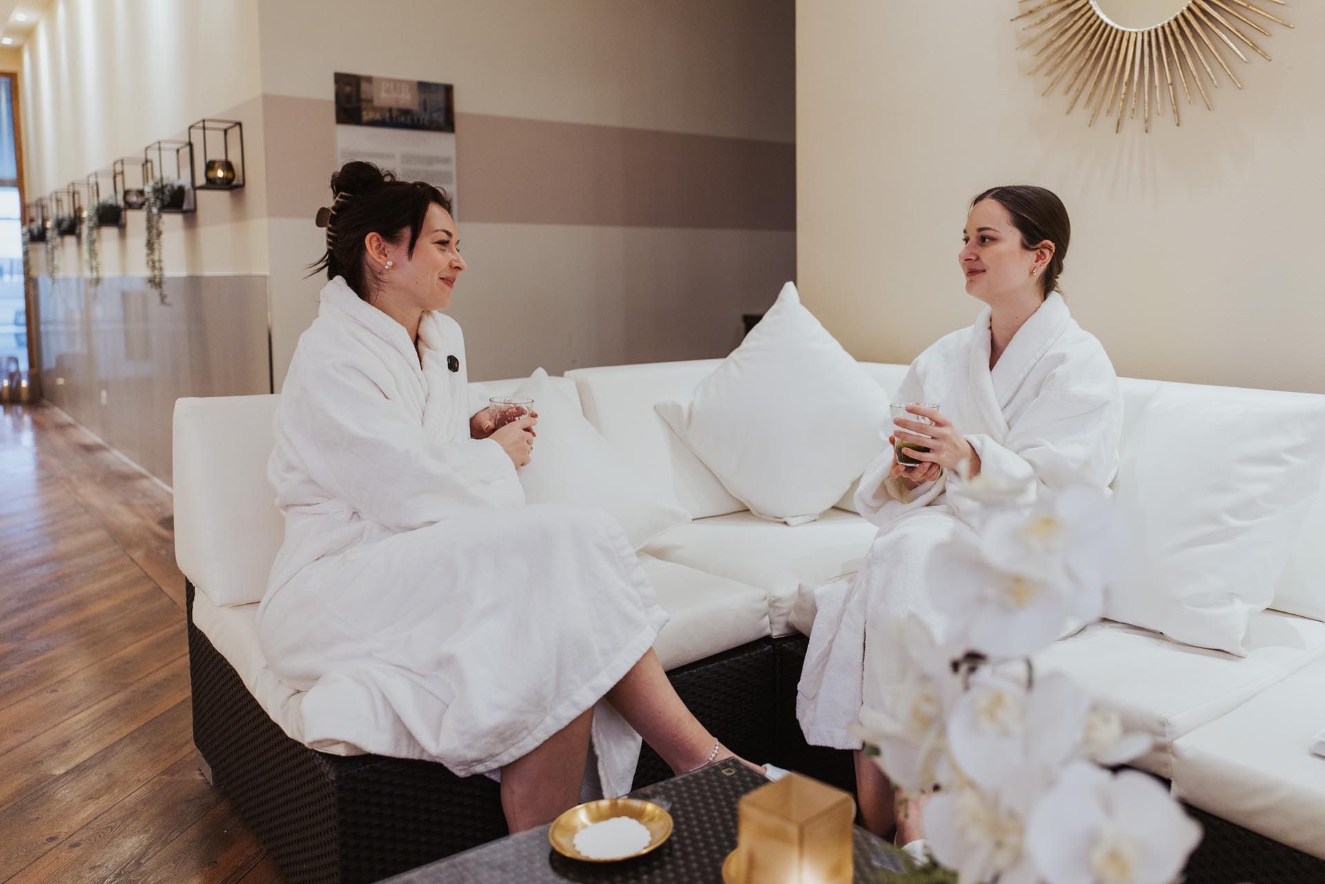 Zwei Frauen in weißen Bademänteln unterhalten sich auf einem weißen Sofa in einer hellen Spa-Lounge.