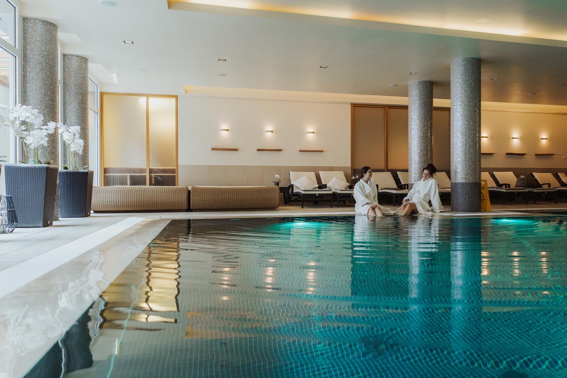 Hotel-Innenpool mit zwei Personen, die am Wasser entspannen, türkisfarbenes Becken und Liegestühle unter warmem Licht