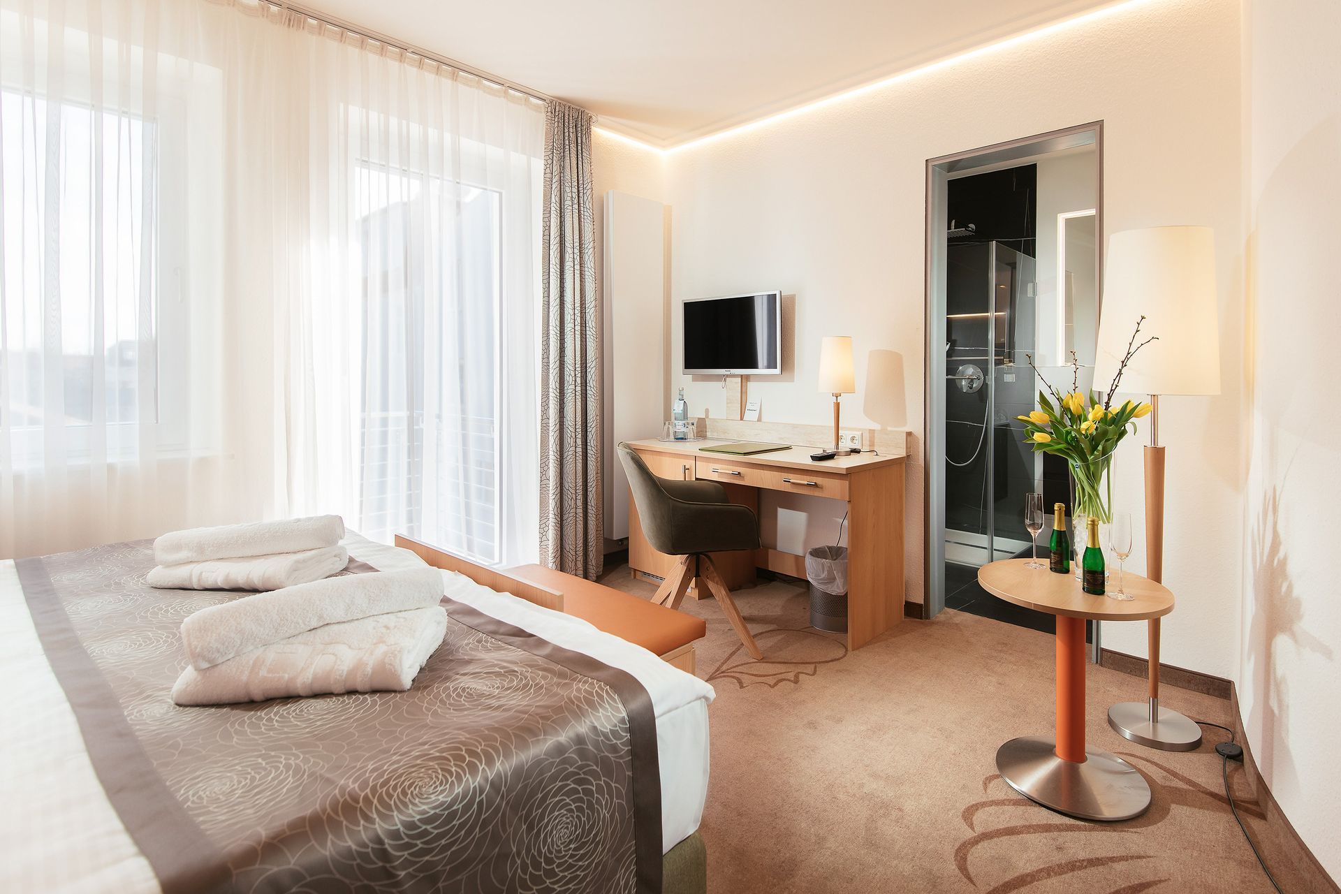 Hotelzimmer mit Bett, Schreibtisch, Fernseher und einem kleinen runden Tisch mit Blumen neben einem Fenster.