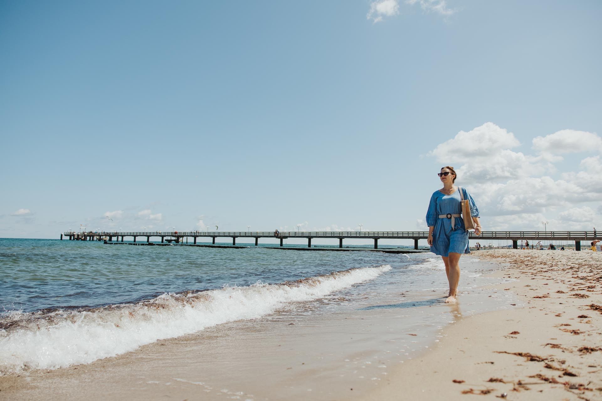 Eine Person in Blau geht an einem Sandstrand in Küstennähe entlang; im Hintergrund befinden sich ein langer Pier und ruhiges Meer.