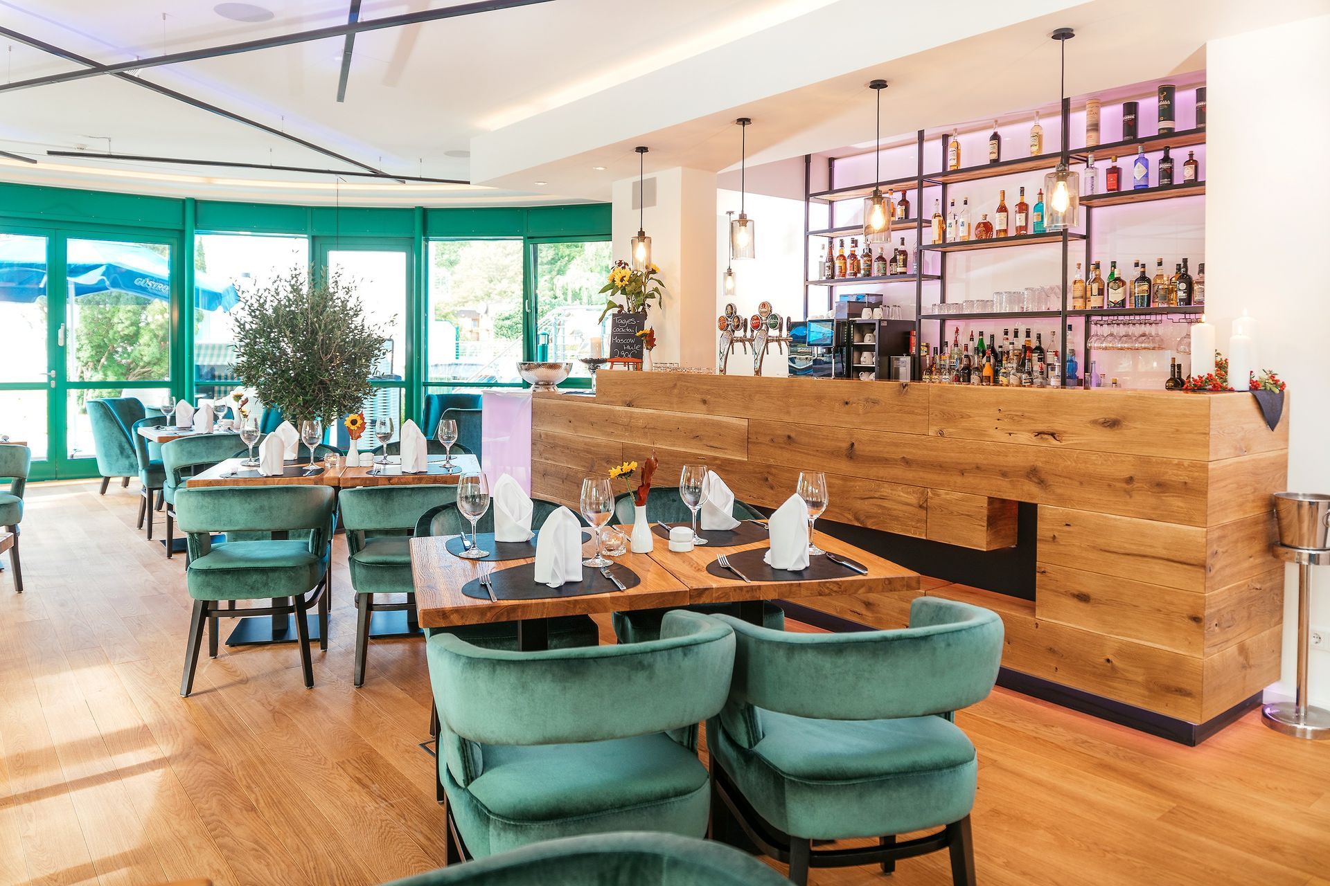 Restaurantinterieur: Holzbar, türkisfarbene Stühle, Tische mit weißen Servietten und große Fenster.