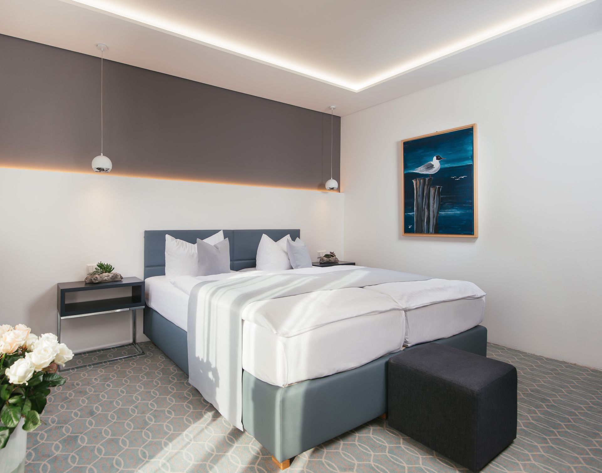 Schlafzimmer mit großem Bett, Nachttischen und einem Gemälde mit einer Möwe. Farbschema in Grau, Weiß und Blau.