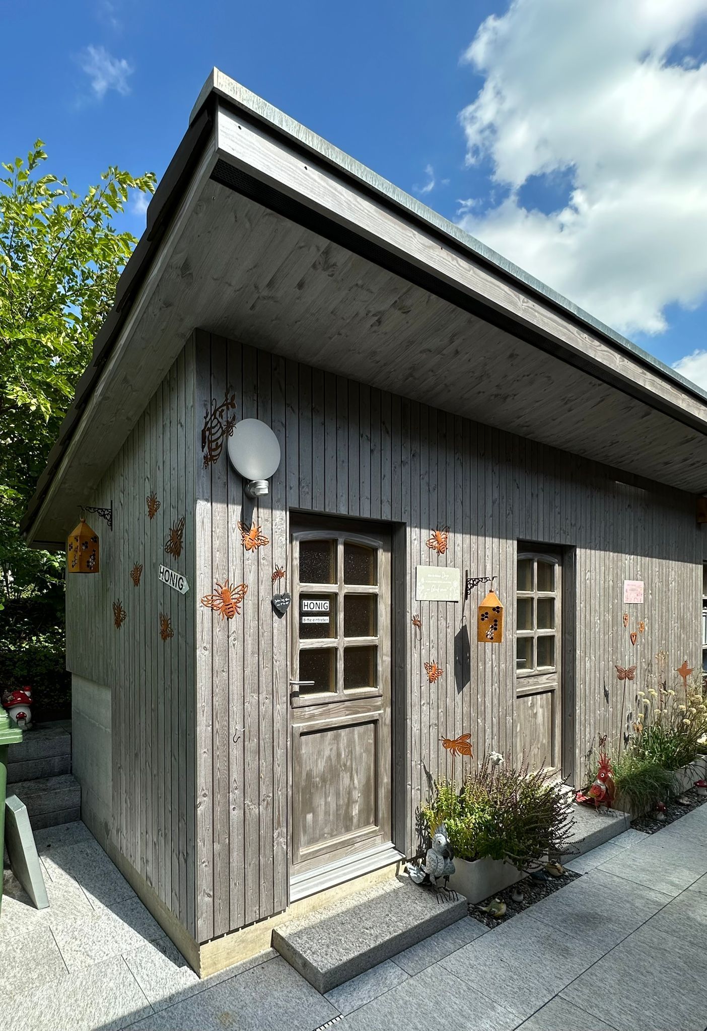 Kleiner, verwitterter Holzschuppen mit zwei Türen, Antenne und Topfpflanzen. Strahlend blauer Himmel.