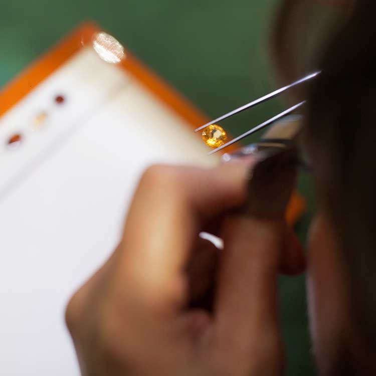 Une personne tient une petite gemme orange à l'aide d'une pince à épiler et l'examine avec une loupe.