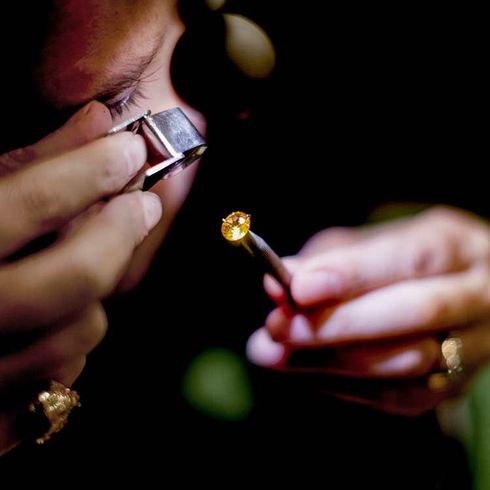 Une personne examine une petite pierre précieuse jaune à l'aide d'une loupe et d'une pince à épiler. Décor sombre.