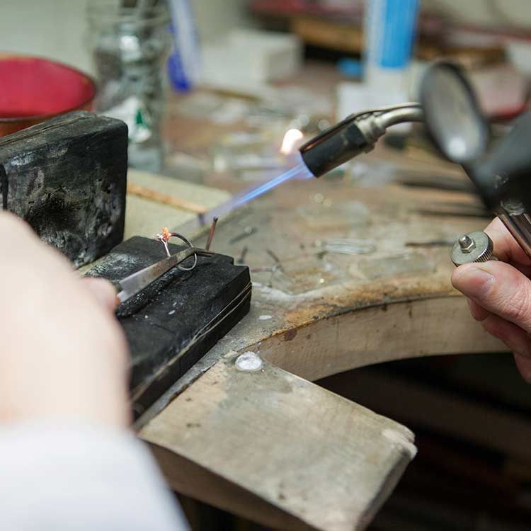 Bijoutier utilisant un chalumeau pour souder du métal sur un établi ; gros plan sur les mains et les outils.