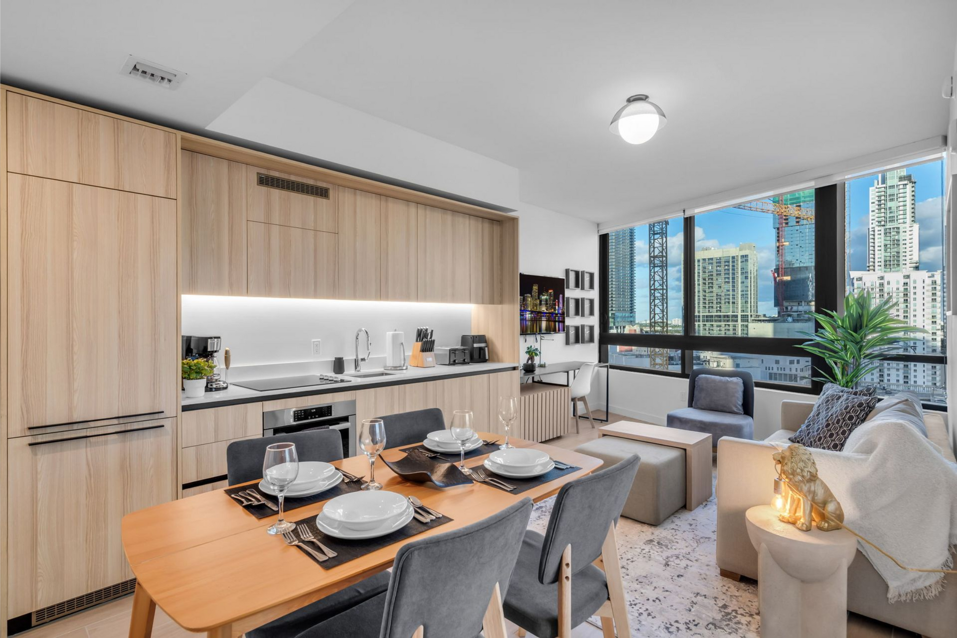Modern open-concept apartment with a kitchen, dining area, and living room; city view through a large window.