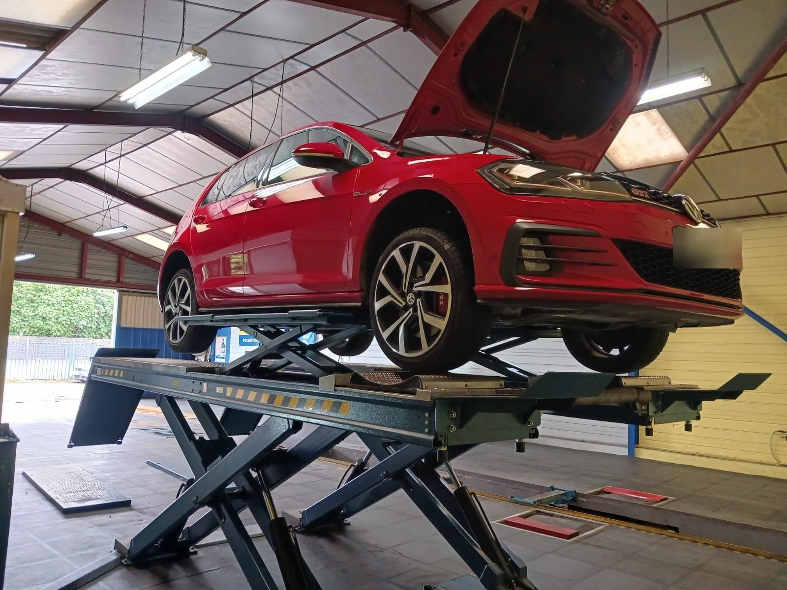 Voiture rouge soulevée sur un pont élévateur à ciseaux dans un garage automobile, capot ouvert.
