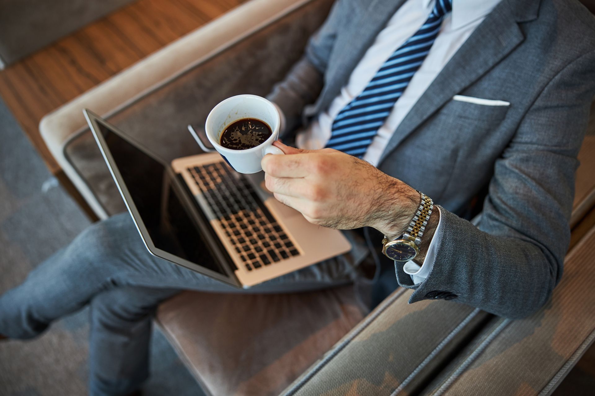 Une personne en costume gris tient une tasse de café tout en utilisant un ordinateur portable.