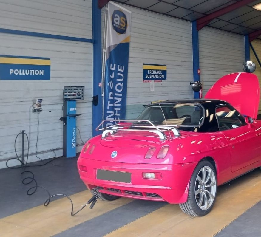 Voiture décapotable rose vif dans un garage, capot ouvert, garée près d'un centre de contrôle des émissions polluantes.