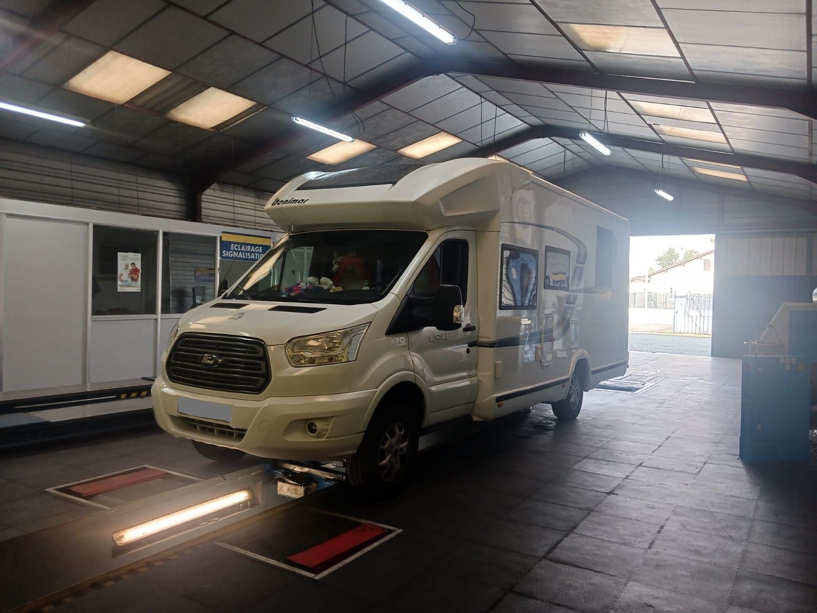 Un camping-car blanc dans un garage avec des lumières vives au plafond et une porte latérale ouverte.