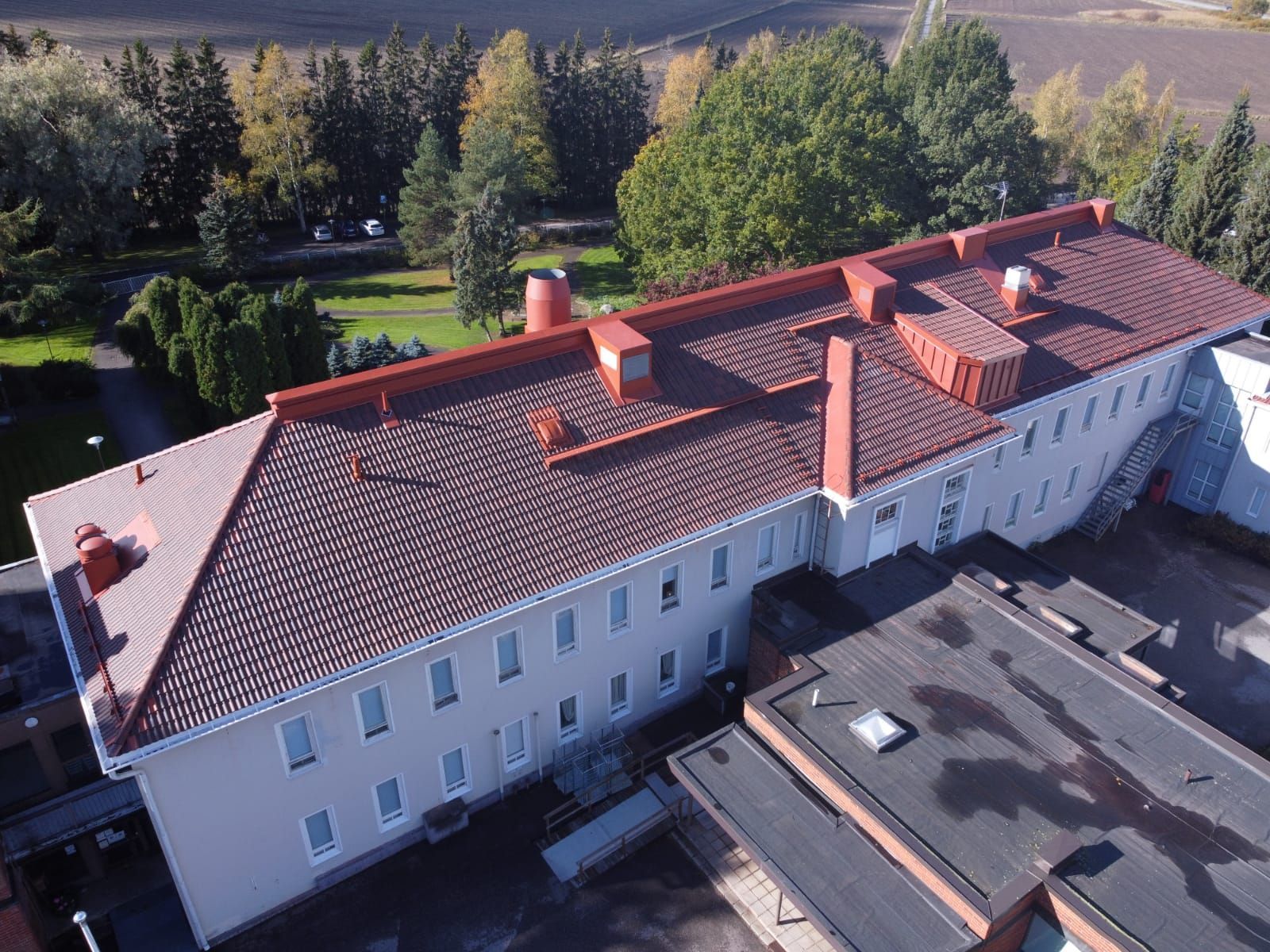 Ilmakuva pitkästä valkoisesta rakennuksesta, jossa on punainen tiilikatto puiden vieressä ja pienempi kattorakennelma.