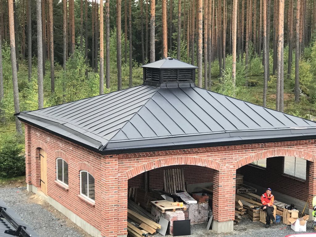 Mustalla peltikatolla varustettu tiilimökki mäntymetsässä, ihmisten työskennellessä avoimen holvikaaren alla.