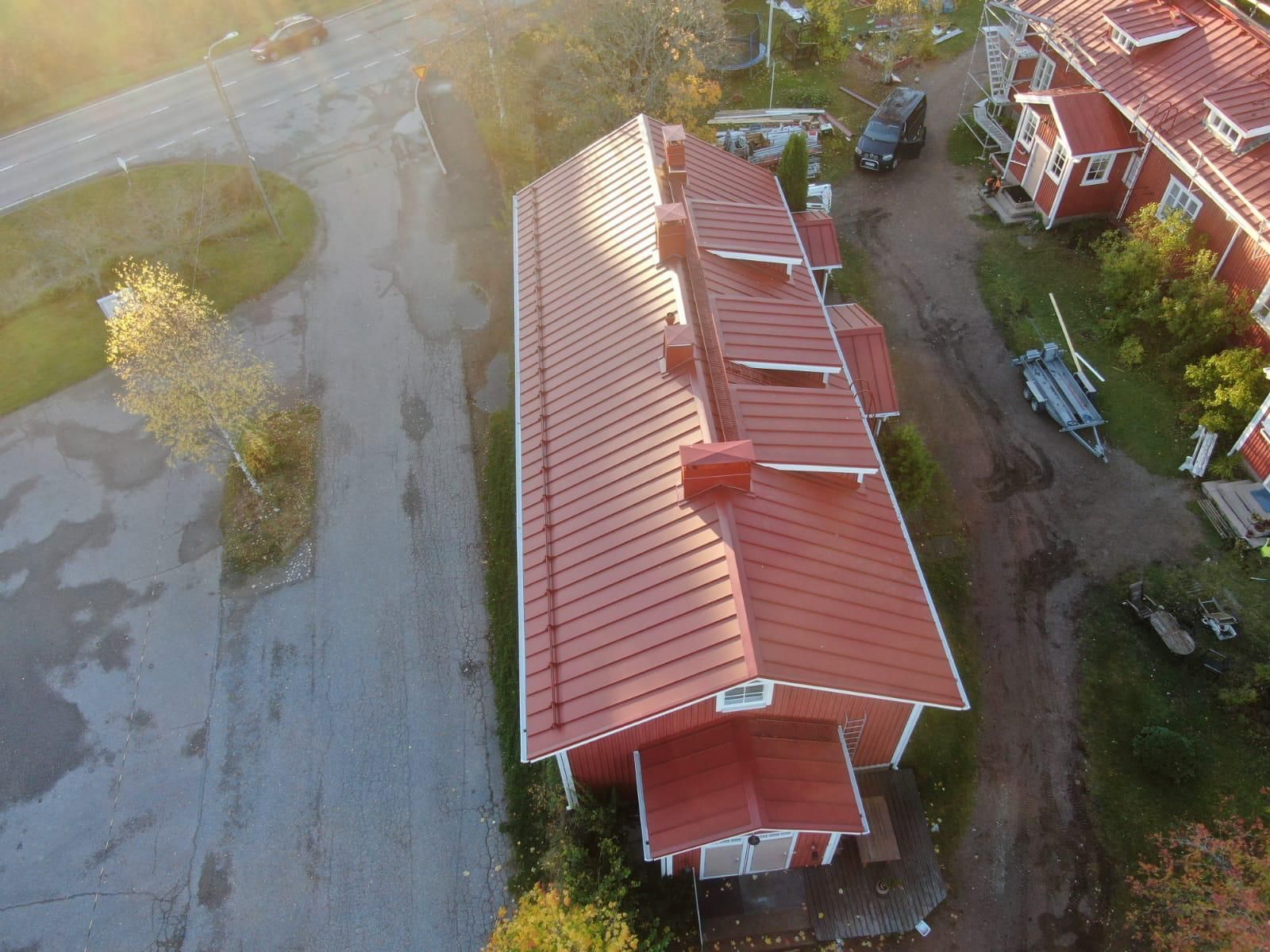 Ilmakuva pitkästä punakattoisesta rakennuksesta tien vieressä ja pysäköidyistä autoista lämpimässä auringonvalossa