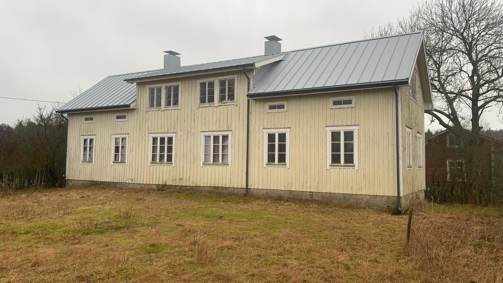 Vanha beigenvärinen puutalo harmaalla metallikatolla nurmikolta päin pilvisen taivaan alla.