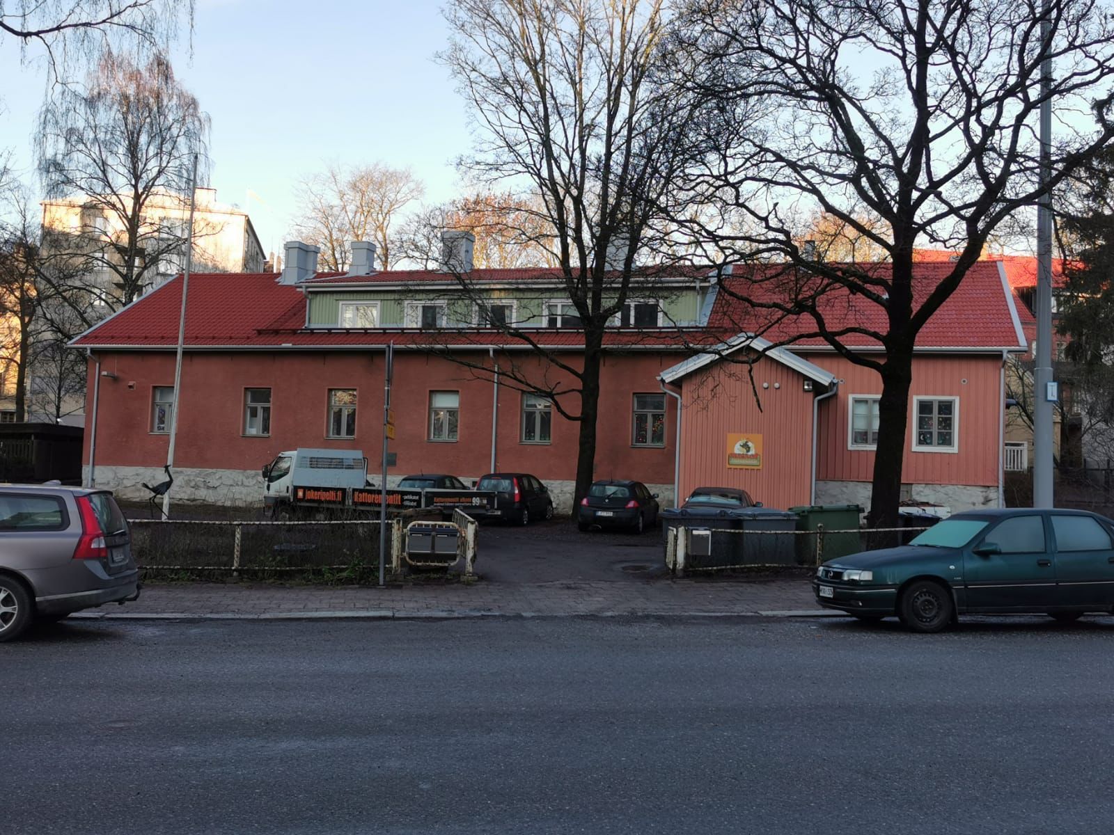 Punainen tiilirakennus, jossa on punainen katto pysäköityjen autojen ja paljaiden puiden takana hämärässä