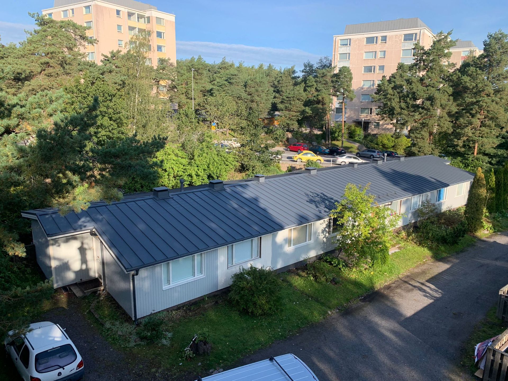 Puiden reunustama sisäpiha, jonka taustalla pitkä harmaakattoinen rakennus ja kerrostalot