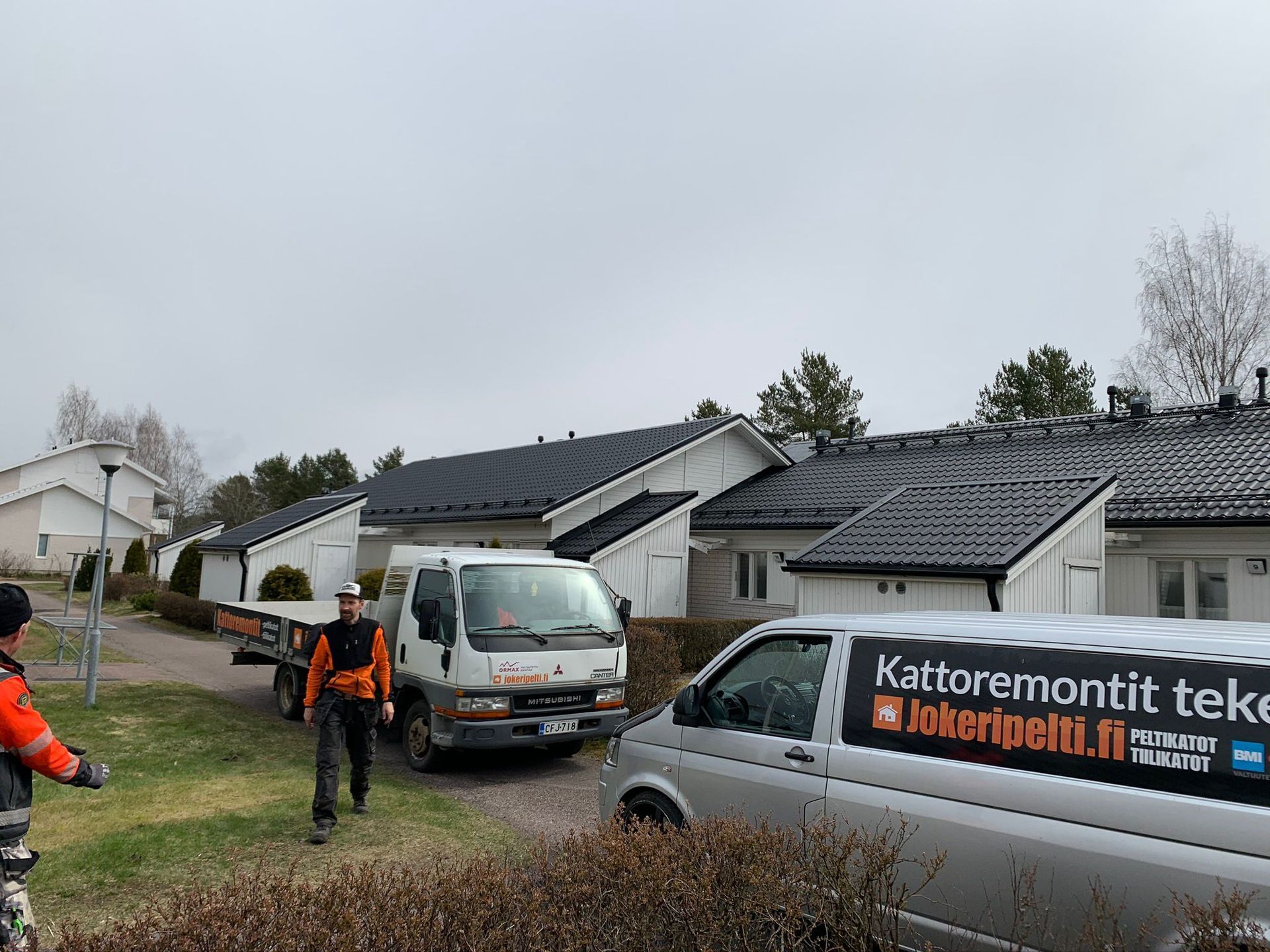 Työntekijöitä ja pakettiautoja rivitalojen ulkopuolella kattokorjauksen aikana pilvisellä alueella