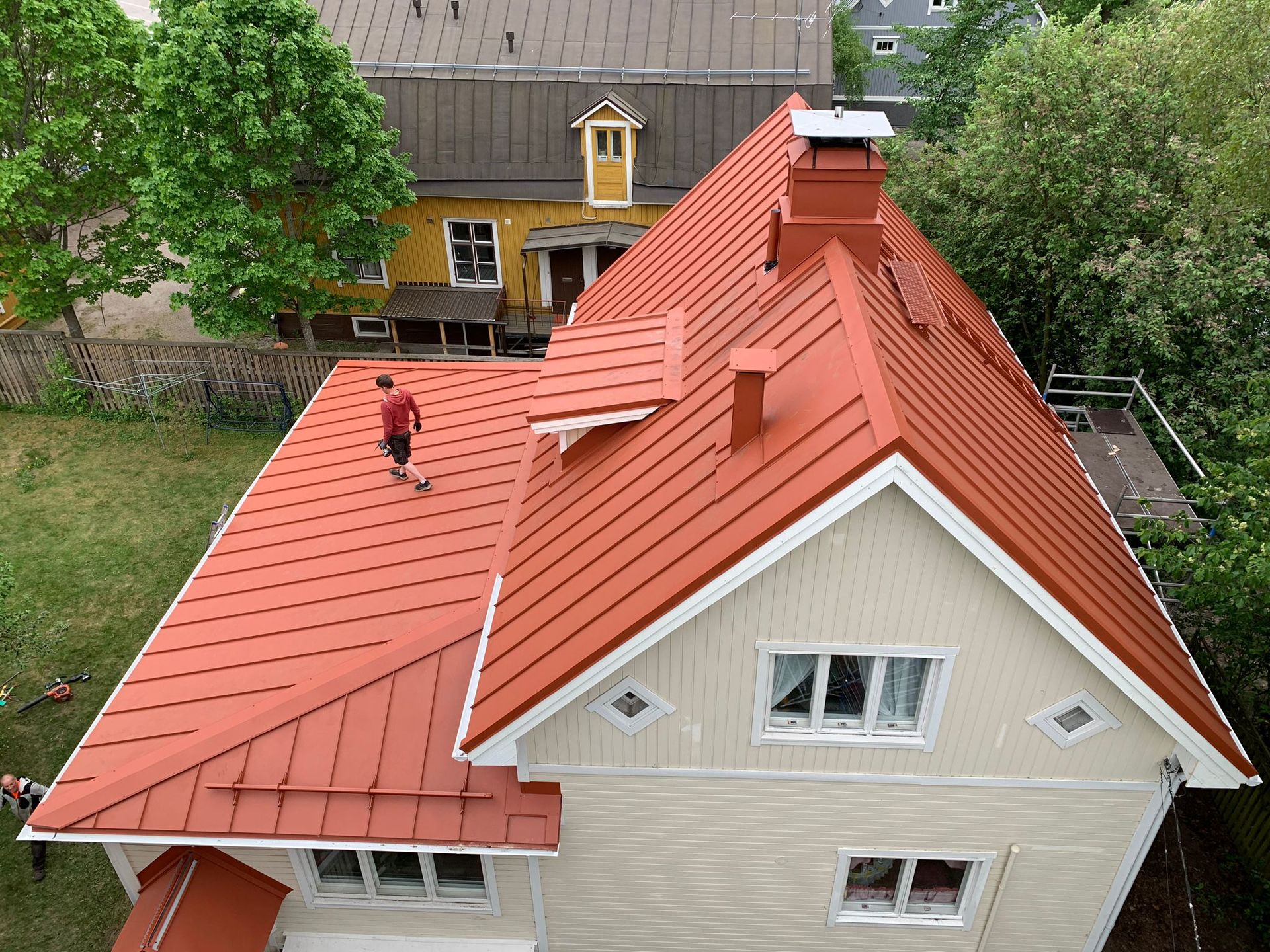 Ilmakuva talosta, jossa on punainen katto ja beigenvärinen ulkoverhous, ja henkilö seisoo katolla.