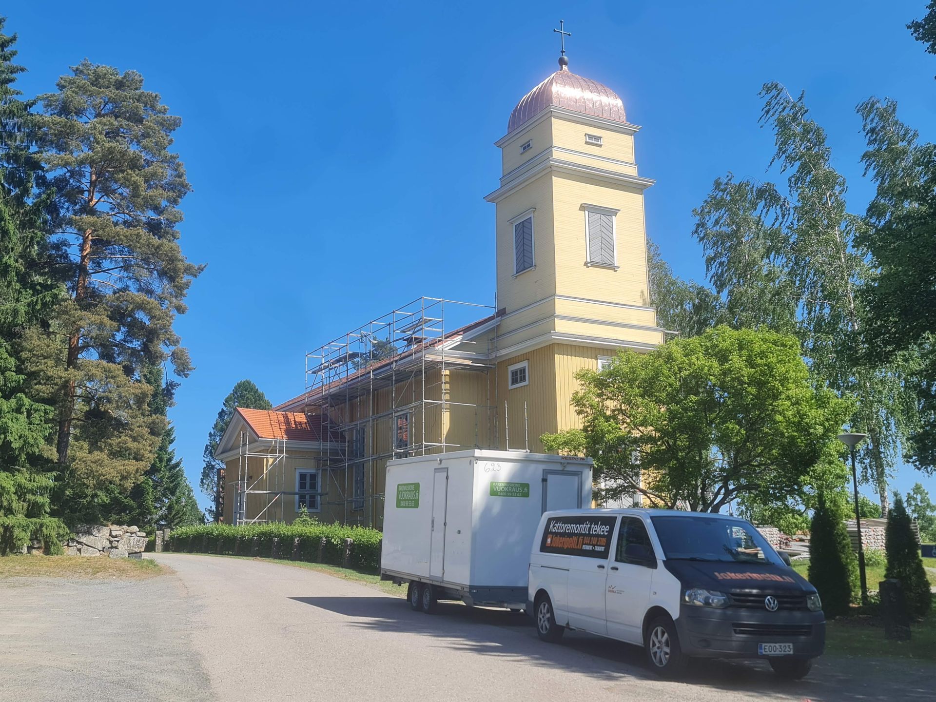Valkoinen hyötykuorma-auto pysäköitynä keltaisen, korkean tornin omaavan kirkon viereen aurinkoisena päivänä