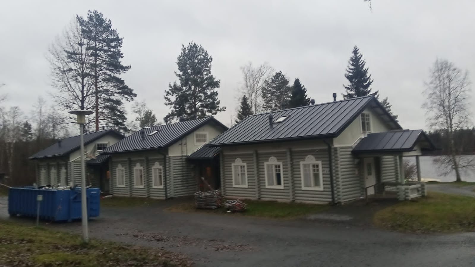 Vanha puinen rakennus, jossa on tumma metallikatto pilvisenä päivänä, sinisten roskisten ja paljaiden puiden vieressä