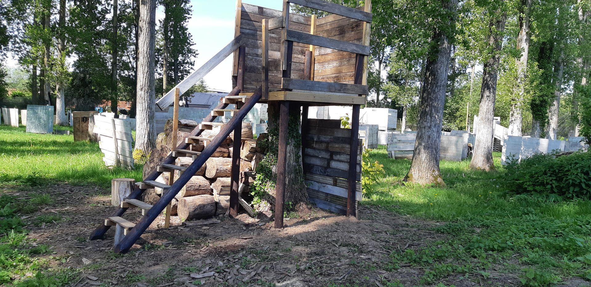 Structure en bois sur un terrain de paintball, entourée d'arbres et de bunkers, avec des escaliers menant à l'étage.