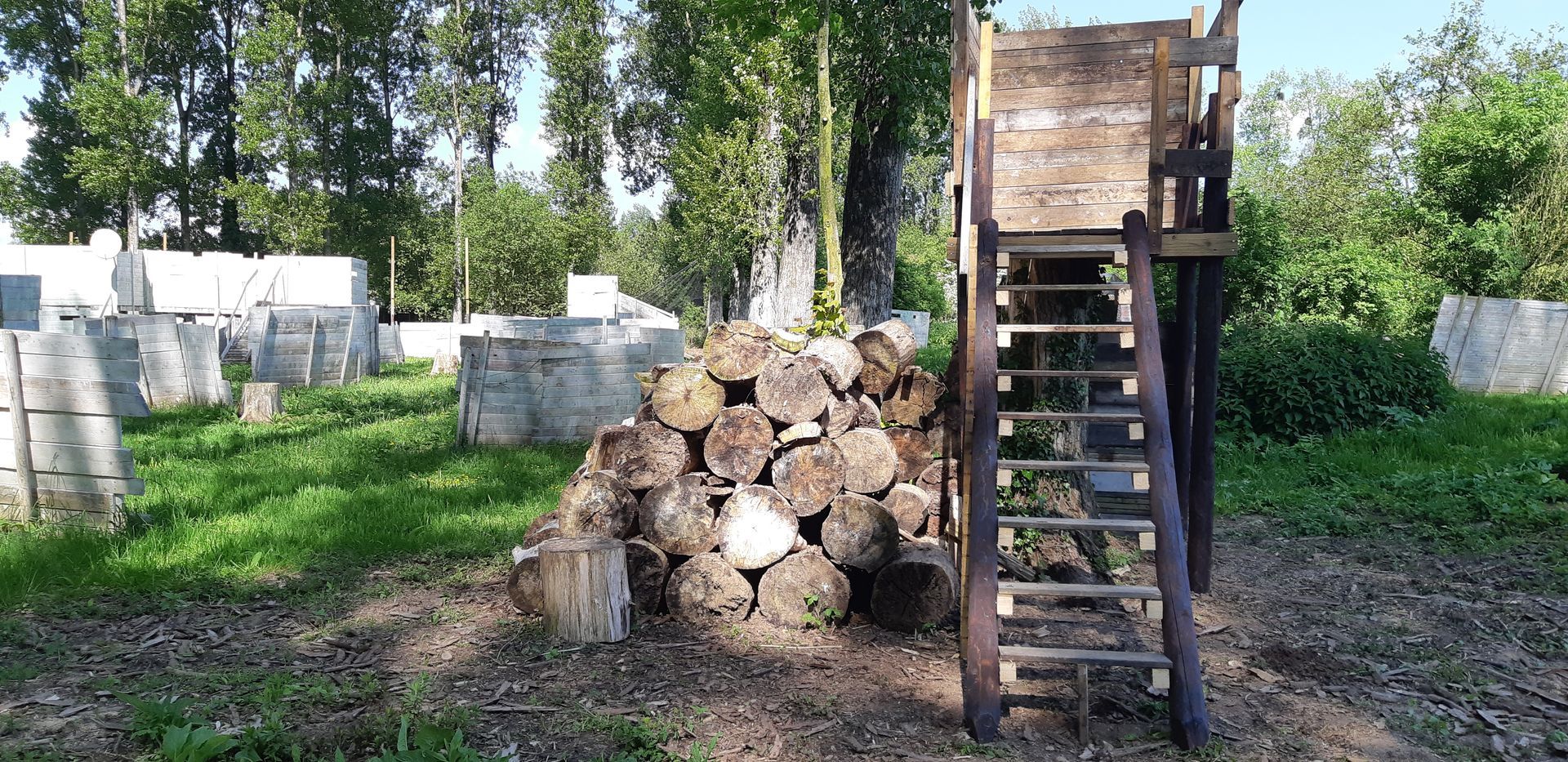Un terrain de paintball avec des bunkers en béton et une tour de guet en bois. Des bûches sont empilées au premier plan. Herbe verte et arbres.