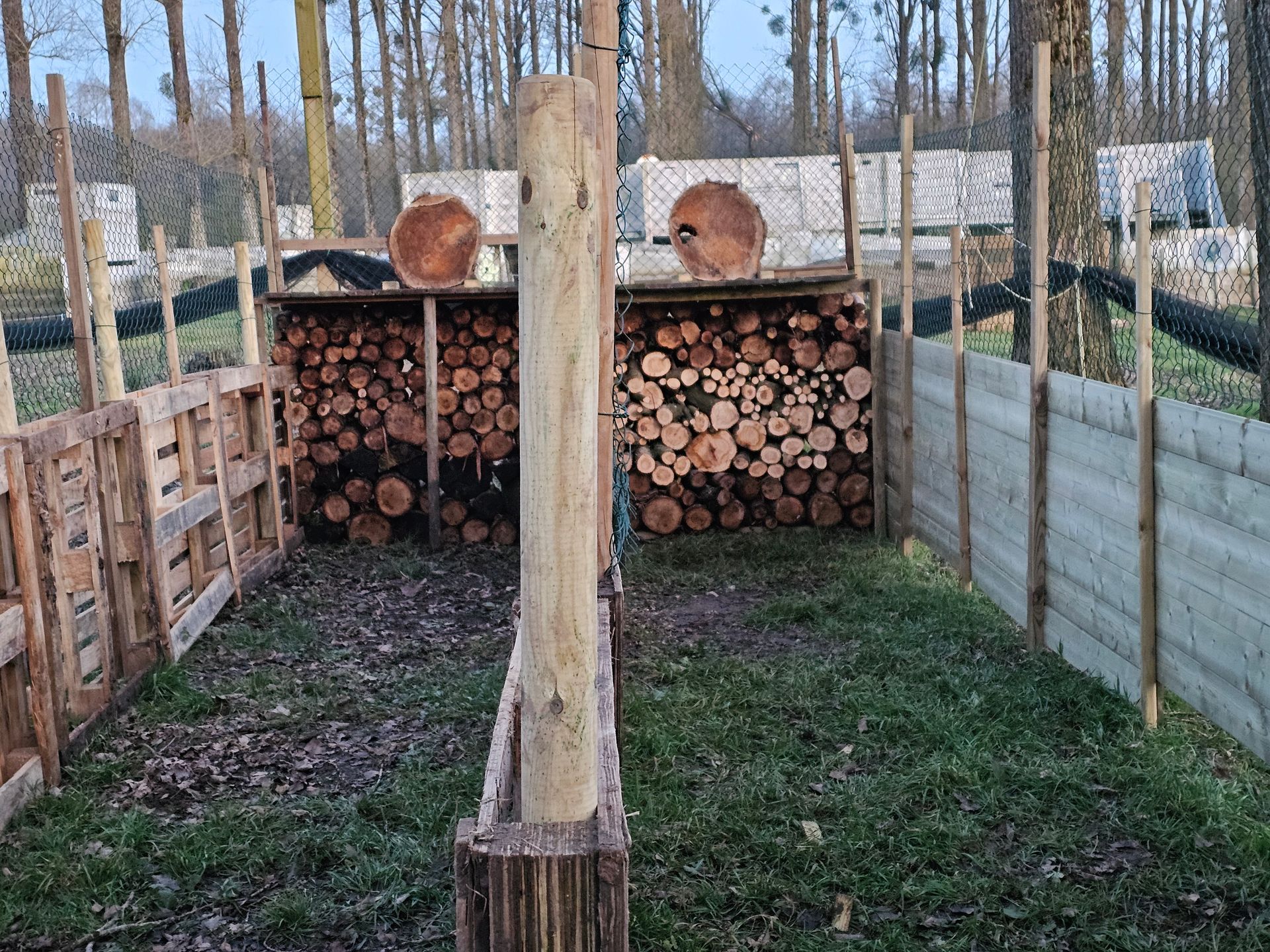 Installation d'un stand de tir à l'arc : deux cibles en bois, un tas de bûches en arrière-plan, des barrières en bois et en filet dans un environnement extérieur.