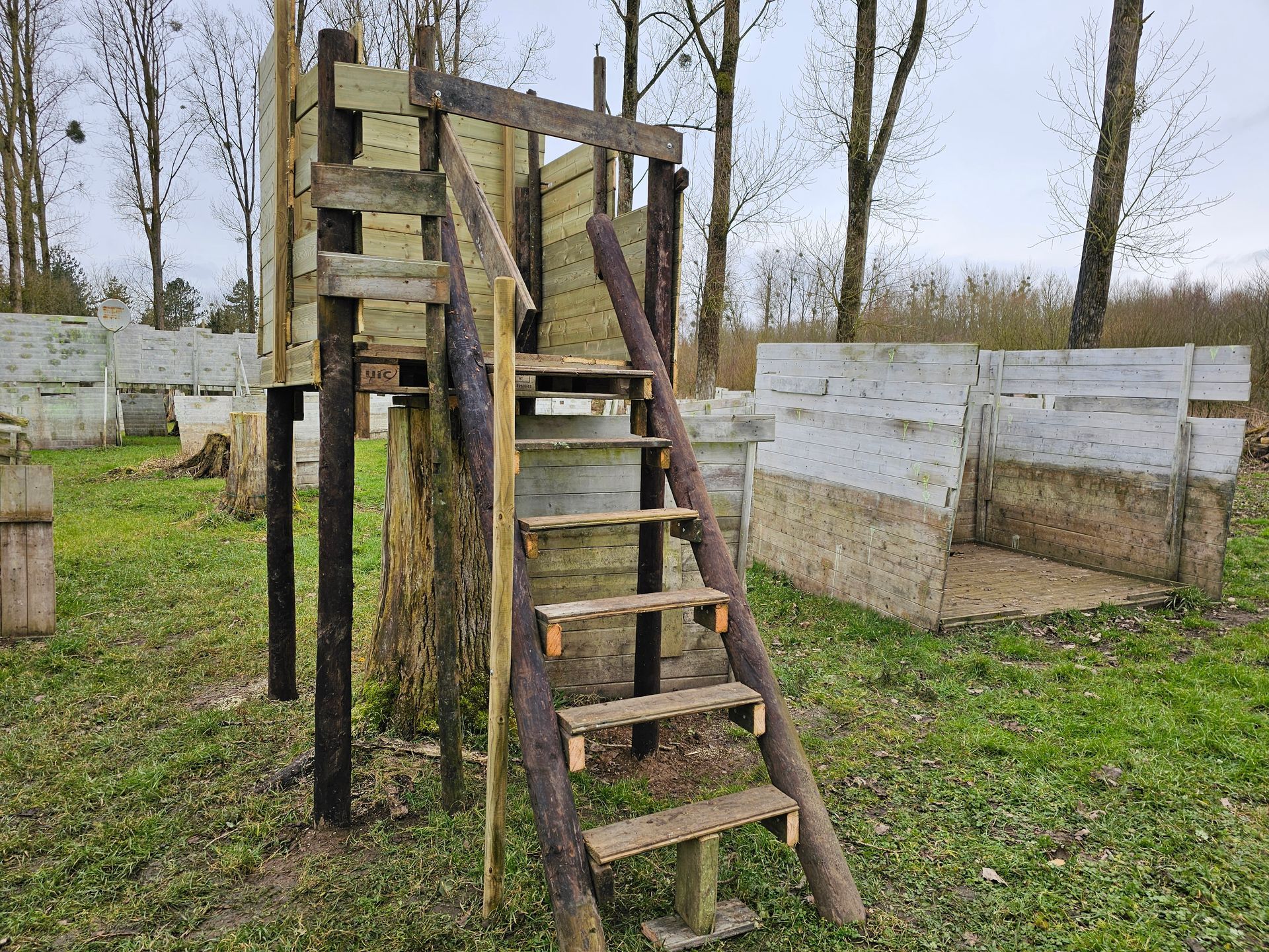 Structure en bois avec escalier dans un terrain de paintball herbeux. Autres obstacles visibles en arrière-plan. Ciel couvert.