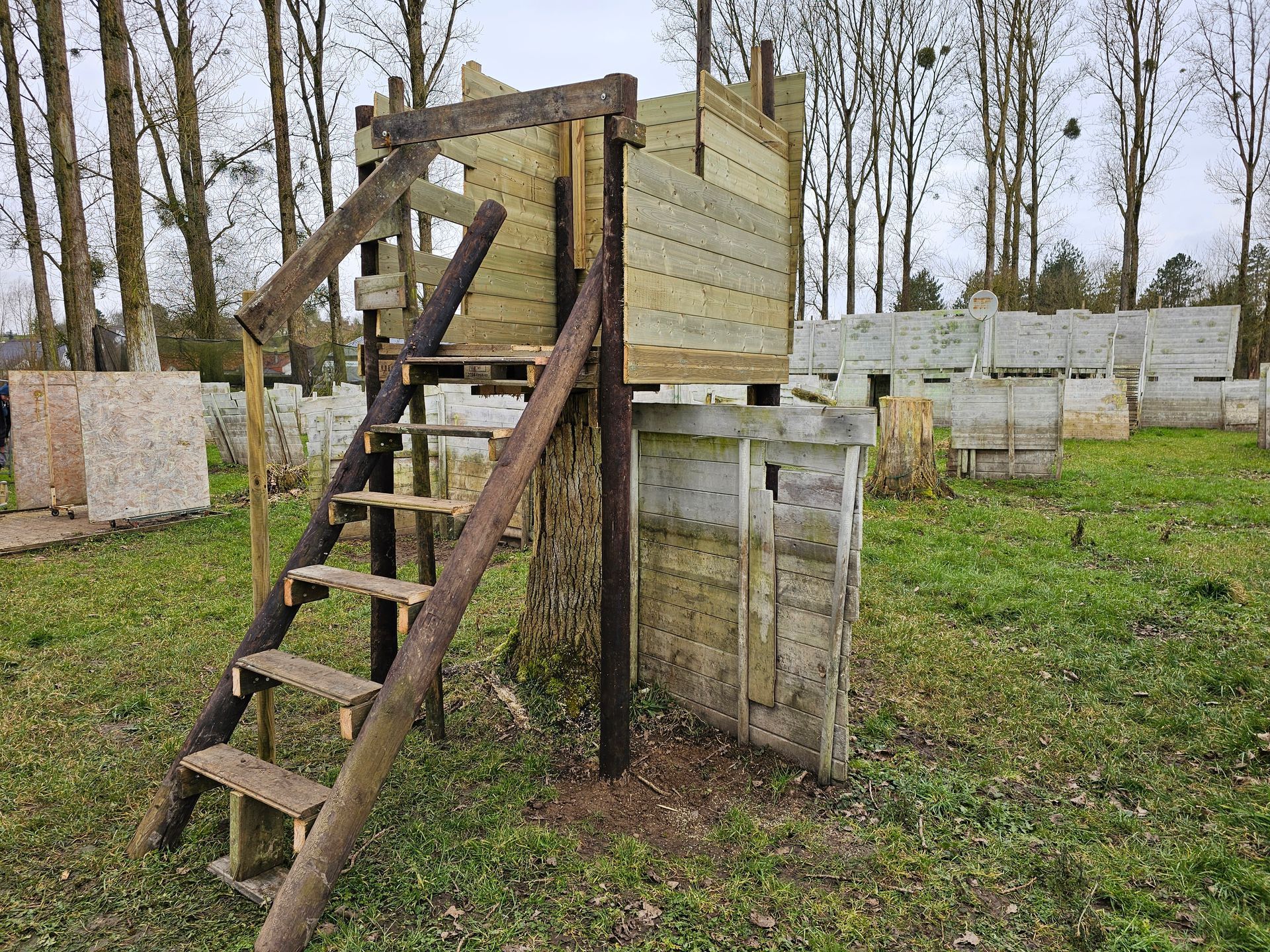 Bunker de paintball en bois avec escalier, situé dans un champ herbeux. Autres bunkers et arbres en arrière-plan.