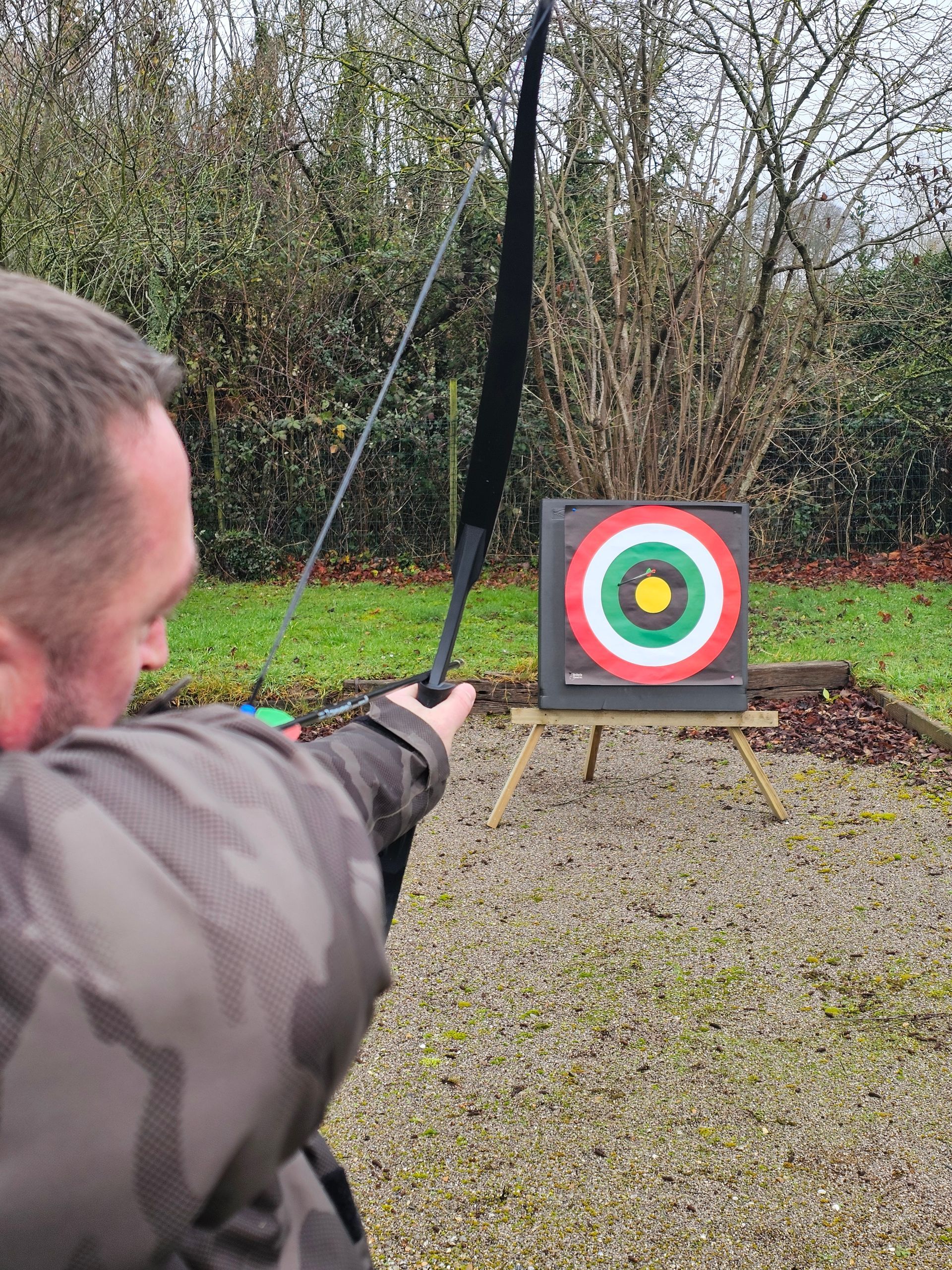 Un homme vise une cible de tir à l'arc avec un arc et des flèches noirs dans un décor extérieur herbeux.