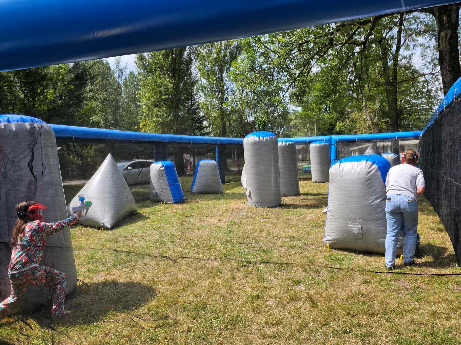 Des personnes jouent au paintball dans un terrain extérieur avec des bunkers gonflables et des arbres en arrière-plan.