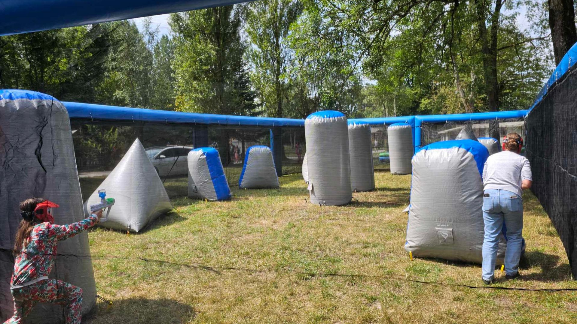 Des personnes jouent au paintball dans un terrain extérieur aménagé avec des bunkers gonflables, des filets de protection et des arbres.