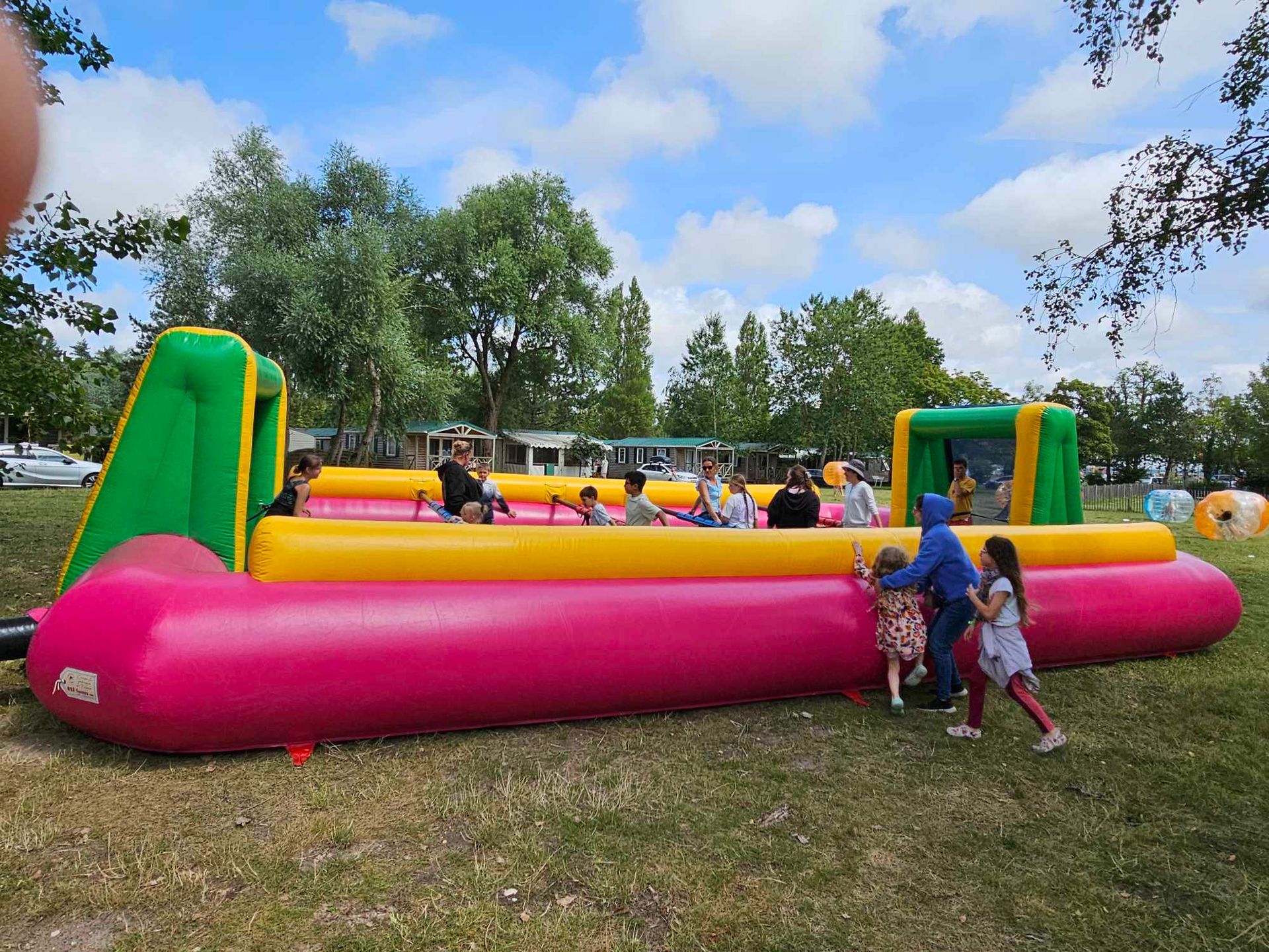 Des enfants jouent sur une grande aire de jeux gonflable avec des sections roses, jaunes et vertes, en plein air sur un terrain herbeux.