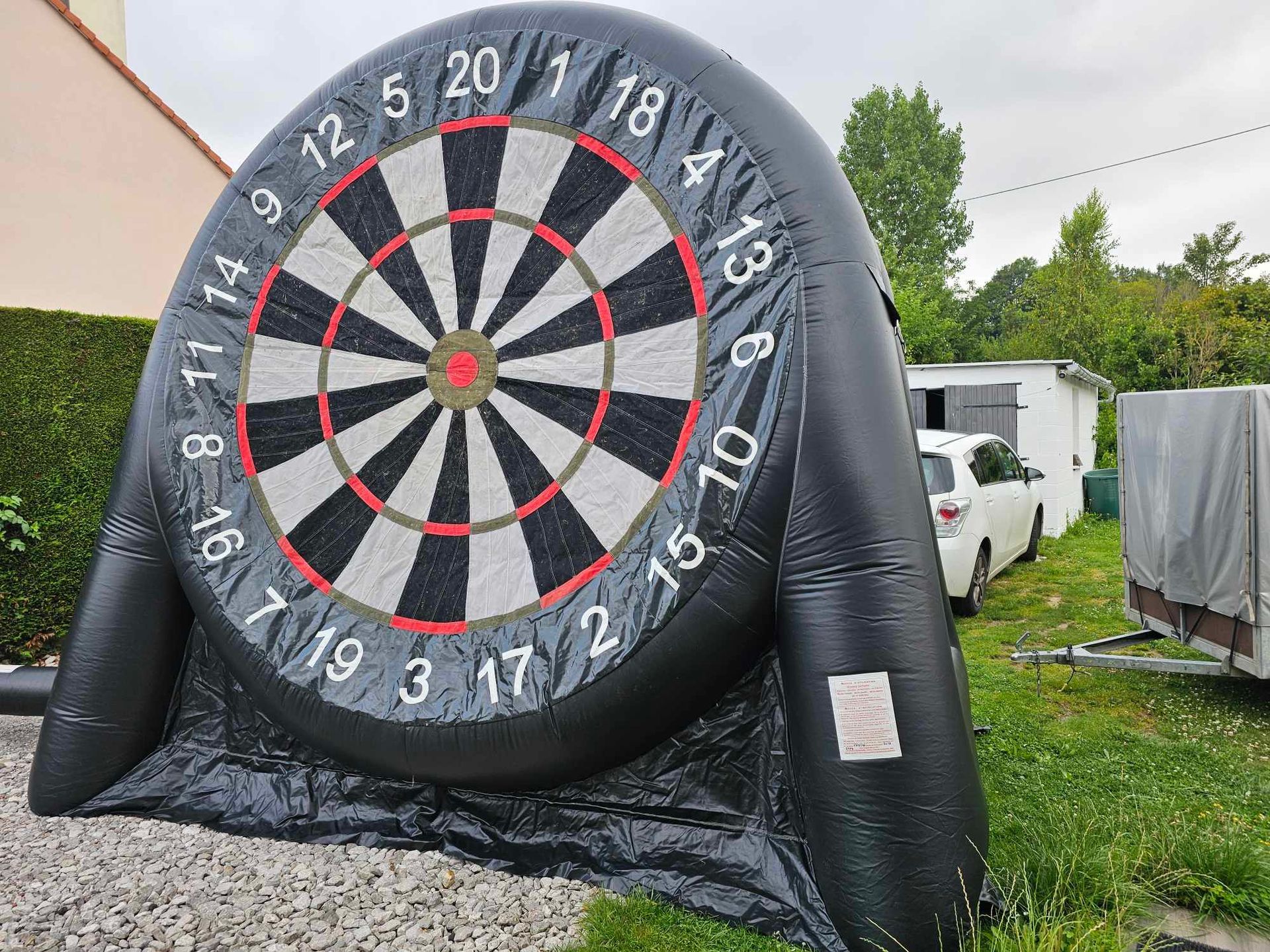 Cible de fléchettes gonflable noire posée sur une surface de gravier à l'extérieur, avec des chiffres, un centre et une voiture blanche en arrière-plan.