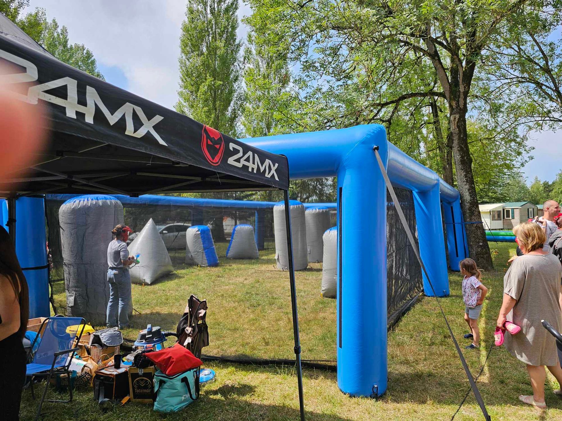 Terrain de paintball avec barrières gonflables. Des personnes se tiennent près des filets et sous un auvent.