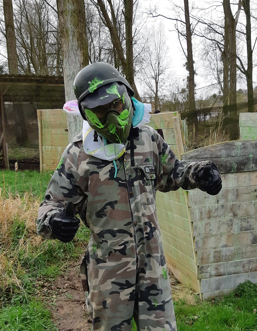 Personne en tenue de paintball camouflage, tenant deux billes de peinture. Porte un casque et un masque. En extérieur.
