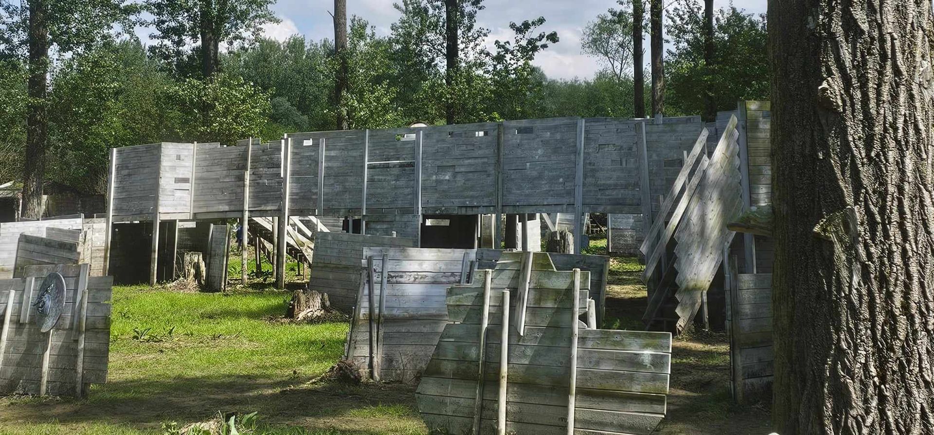 Structures en bois sur un terrain de paintball, entourées d'arbres et d'herbe, par une journée ensoleillée.