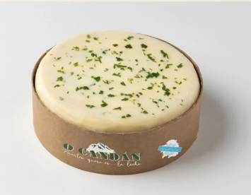 Queso redondo en recipiente de cartón marrón; adornado con hierbas verdes.