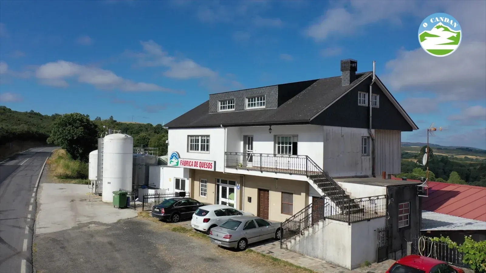 Edificio blanco de dos pisos con techo oscuro y un cartel, con autos estacionados al frente, contra un cielo brillante.