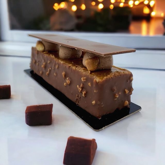 Un dessert rectangulaire au chocolat, garni de mousse et d'éclats de chocolat, présenté sur un plateau blanc parsemé de cubes.