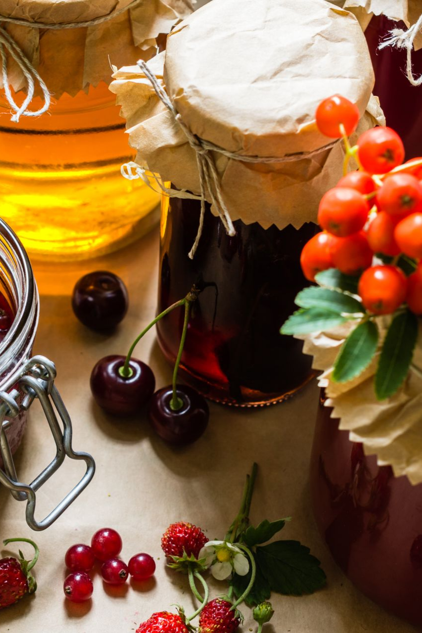 Des pots de confitures colorées et de miel sur une table, décorés de cerises fraîches, de fraises des bois et de baies de sorbier.